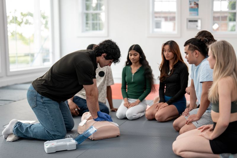 An instructor performs CPR on a mannequin while a group of ethnically diverse students attentively observes during a training session indoors, promoting health, teamwork, and first aid education.