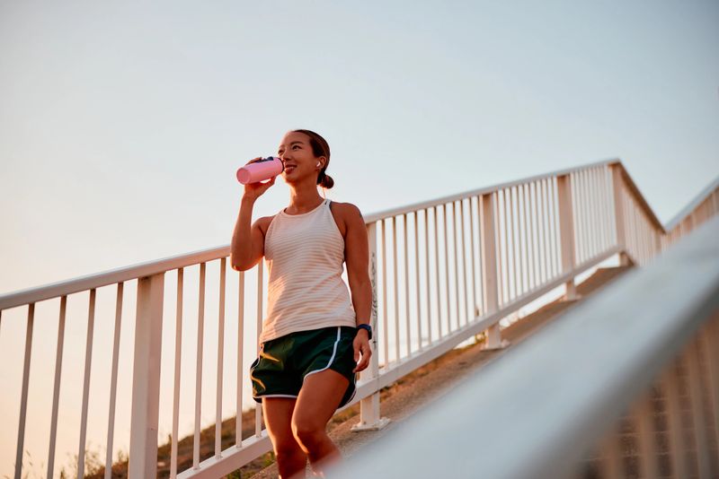 Young woman sipping from her pink water bottle while climbing stairs during an outdoor workout, embodying determination and vitality in her fitness journey