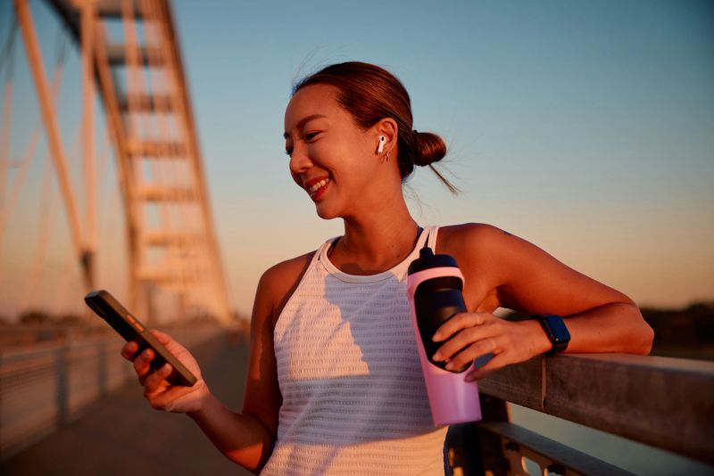 Young sportswoman smiling while using her smartphone after an invigorating training session on the bridge during a stunning sunset, embracing a healthy and active lifestyle