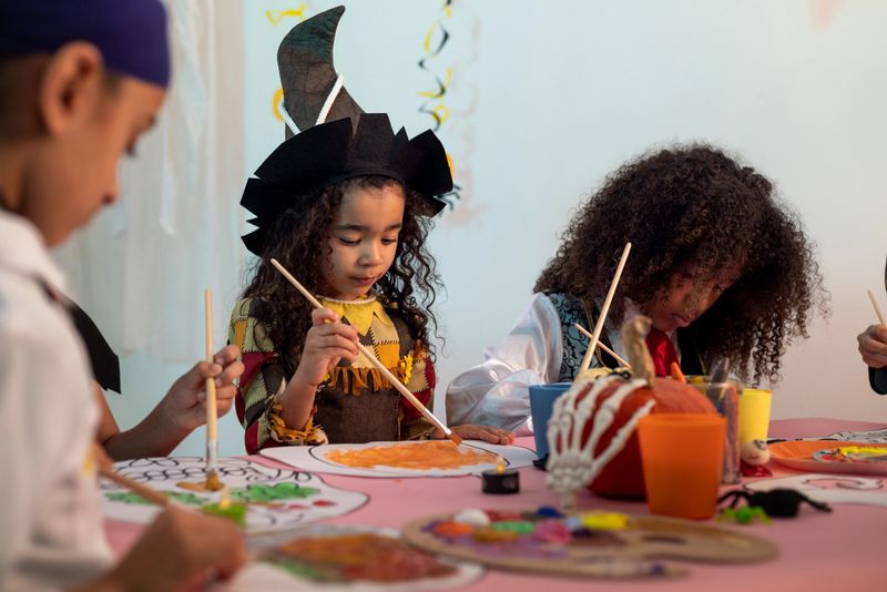 A wide shot of children dressed in various Halloween costumes are painting spooky Halloween artwork with colourful paints. The room and table are decorated with Halloween decorations such as spiders and pumpkins.Videos are available similar to this scenario.
