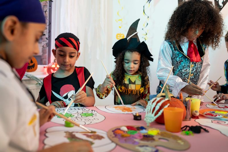 A wide shot of a Halloween arts and crafts table decorated with pumpkins, toy spiders, and paints. The setup includes colourful paints on a pumpkin-shaped plate, paintbrushes in cups, crayons, and paper with Halloween-shaped items outlines ready for colouring. Children, dressed in fancy dress, are painting Halloween images.Videos are available similar to this scenario.
