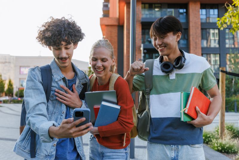 Three diverse students engage in a lighthearted moment of sharing while holding books and a phone, standing outdoors.