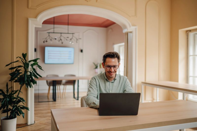 Young adult businessman smiling while working on his laptop in a bright, modern office, exuding confidence and professionalism