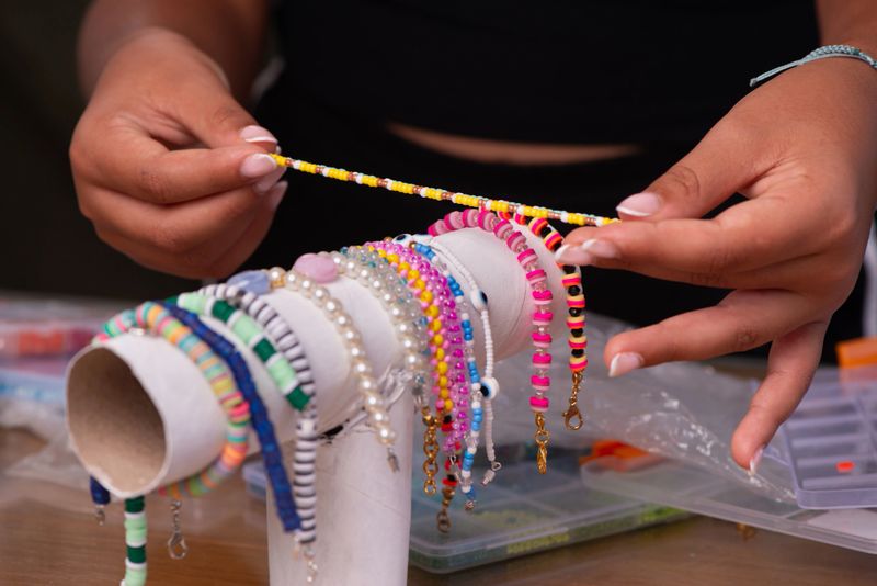 Close-up of hands holding a delicate yellow beaded bracelet above a display stand filled with multiple colorful handmade bracelets. The workspace is surrounded by organized bead boxes, highlighting creativity, jewelry design, and the process of crafting unique accessories.