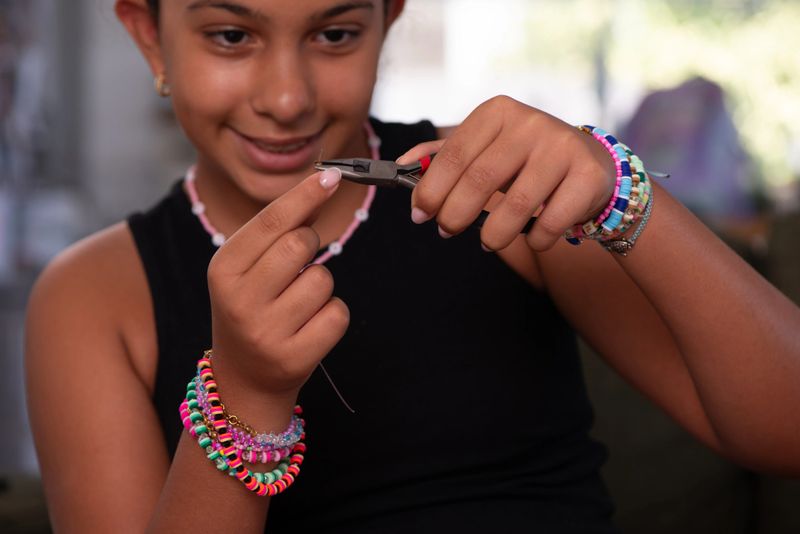 Close-up of a smiling girl using jewelry pliers to work on a piece of handmade jewelry. She wears multiple colorful beaded bracelets on her wrists, showcasing creativity, craftsmanship, and the joy of do-it-yourself jewelry making.