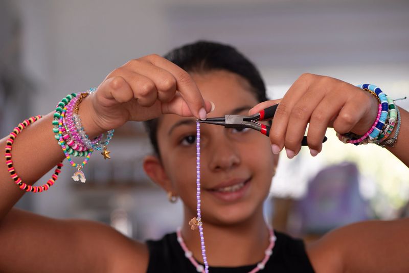 Close-up of a girl smiling while using pliers to work on a string of purple beads. She wears several colorful handmade bracelets on her wrists, highlighting creativity, craftsmanship, and the process of do-it-yourself jewelry making.