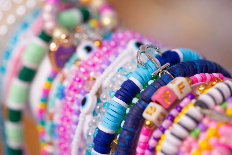 A detailed macro shot of vibrant handmade beaded bracelets arranged together, featuring a variety of colors, textures, and charms, including an anchor and lettered beads. The image highlights creativity, craftsmanship, and the beauty of artisanal jewelry design.