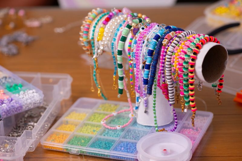 A close-up of a collection of colorful handmade beaded bracelets displayed on a simple stand, surrounded by organized boxes of small beads in various shades. The vibrant jewelry pieces showcase different designs, textures, and patterns, highlighting creativity, craftsmanship, and the art of DIY accessory making.