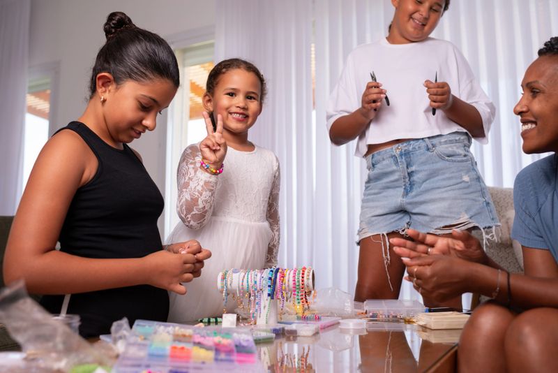 A joyful family moment as children and a woman create handmade beaded jewelry together at home. One smiling girl shows a peace sign while others focus on crafting, surrounded by colorful beads, bracelets, and tools on the table. The scene captures creativity, bonding, and happiness through a fun DIY activity.