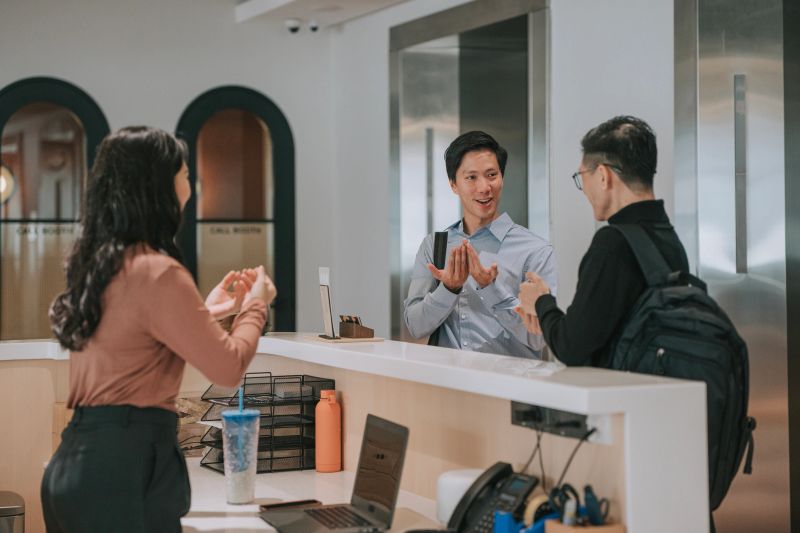 Asian colleagues engaging in conversation using sign language at a workplace reception area, highlighting inclusive communication in a modern office environment. Kuala Lumpur, Malaysia.