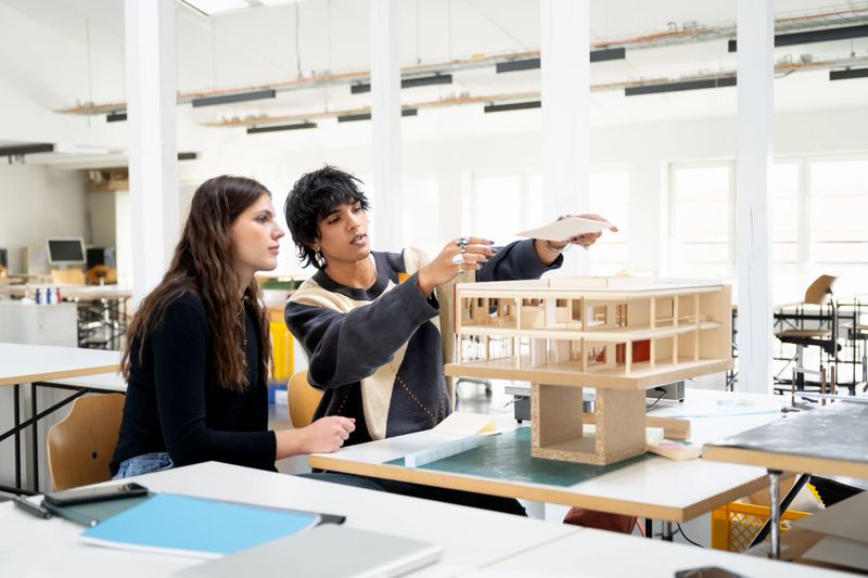 A male and female student work closely together on a wooden architectural model. The male student adjusts an element while explaining, and the female student observes attentively, showing active teamwork and design discussion.