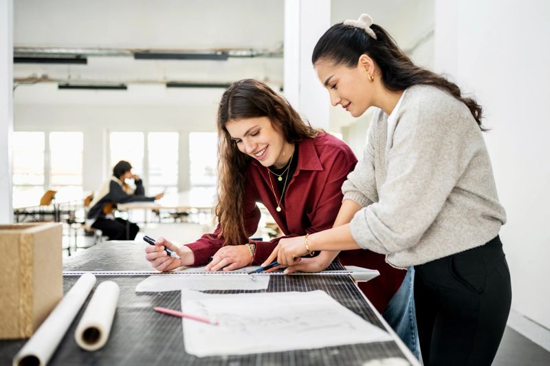 Two students work side by side, smiling as they measure and sketch designs. Their teamwork highlights the collaborative nature of architectural education in the studio.