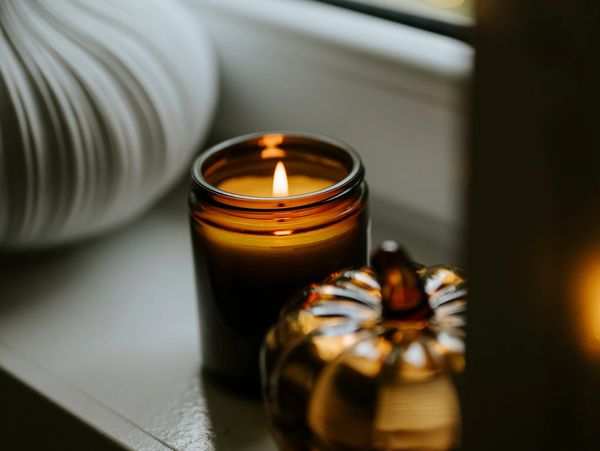 A lit candle in a brown jar next to a decorative pumpkin on a windowsill.