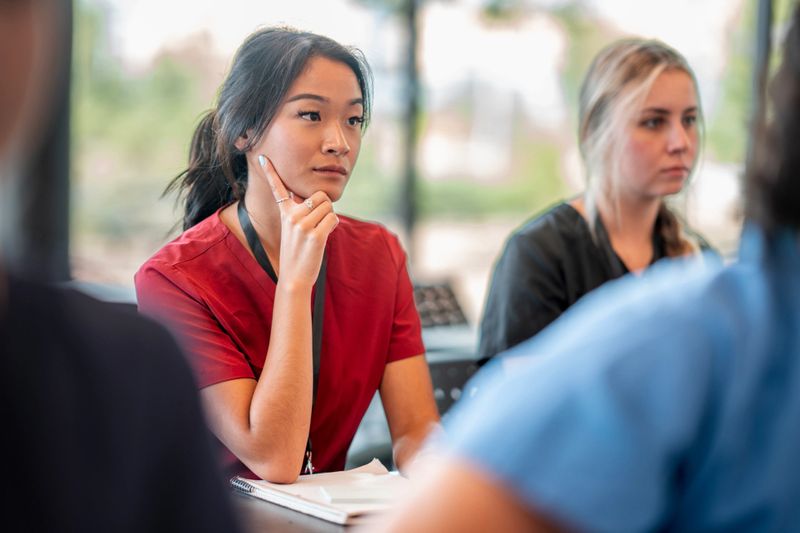 A young nurse in training, wearing a red uniform, attentively participates in an academic session with peers, showcasing focus and determination in a professional learning environment.