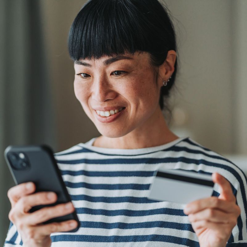 Smiling asian woman holding a credit card while using her smart phone, enjoying the convenience of making an online purchase from the comfort of her home. Embracing modern e-commerce