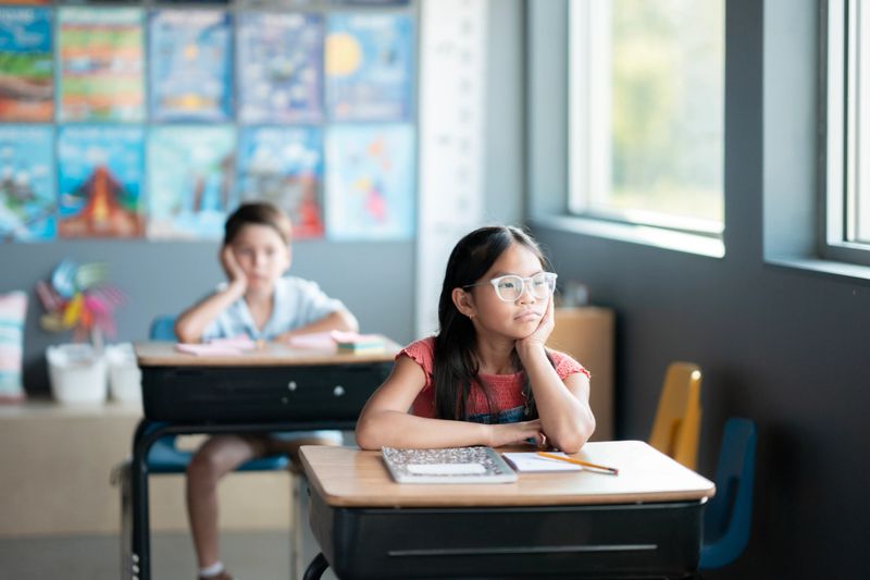 A young girl with glasses sits thoughtfully at her desk in a bright elementary classroom, with a boy sitting in the background, indicating an educational and engaging school environment.