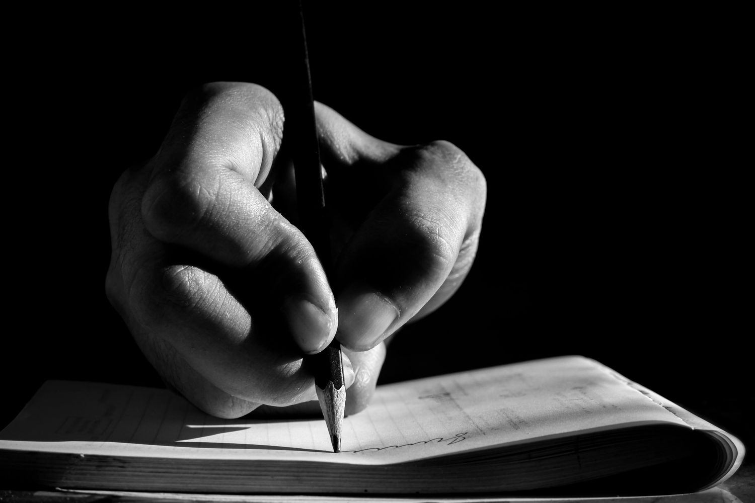 Close-up of a hand writing in a notebook with a pencil in black and white.