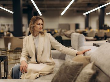 Woman in beige coat sitting and touching a sofa in a furniture store.