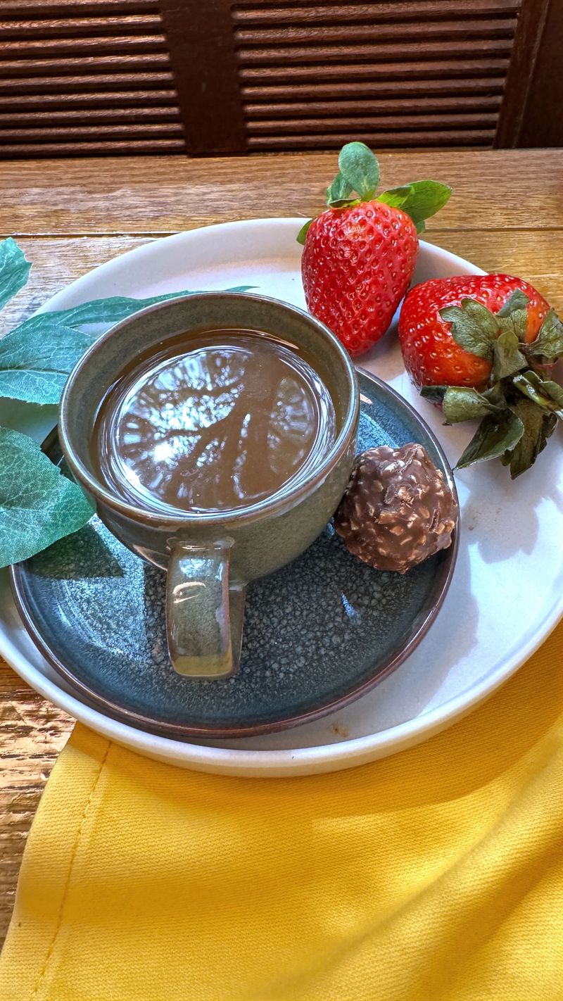 Steaming cup of coffee sits on saucer with fresh strawberries and chocolate treat, creating cozy and inviting breakfast scene on rustic wooden table with yellow napkin
