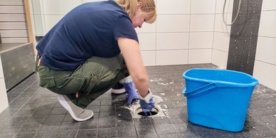 Woman cleaning a shower drain wearing gloves with a blue bucket nearby.