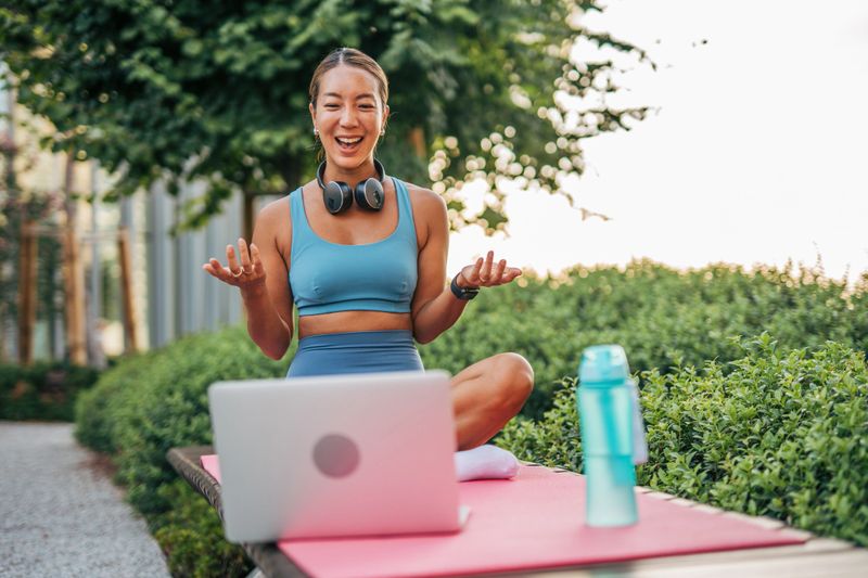 Young asian fitness instructor leading an online yoga class in a park, sitting in lotus position on a yoga mat with a laptop and headphones, promoting healthy lifestyle and wellness