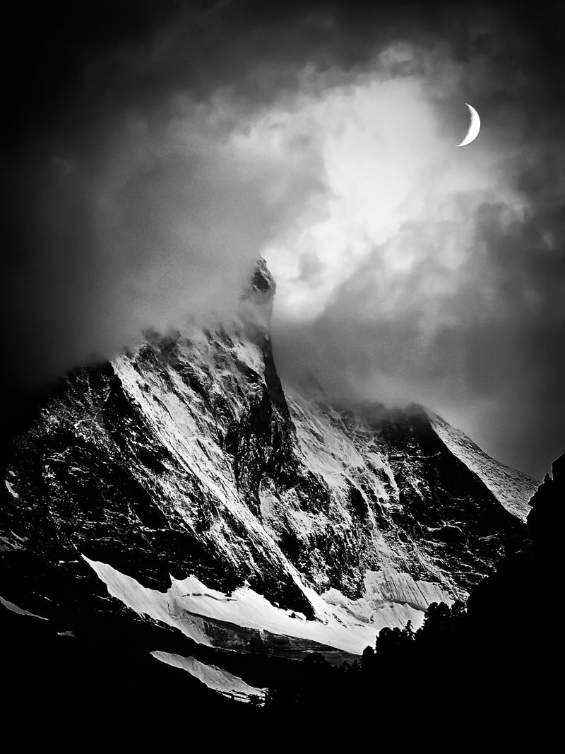 Black and white photograph of the Matterhorn in Zermatt, Switzerland. The dramatic scene captures the iconic peak partially shrouded in clouds, with a crescent moon glowing through the mist above the mountain. The snow-covered slopes stand out starkly against the dark shadows, emphasizing the rugged alpine landscape and the raw atmosphere of the high-altitude environment.