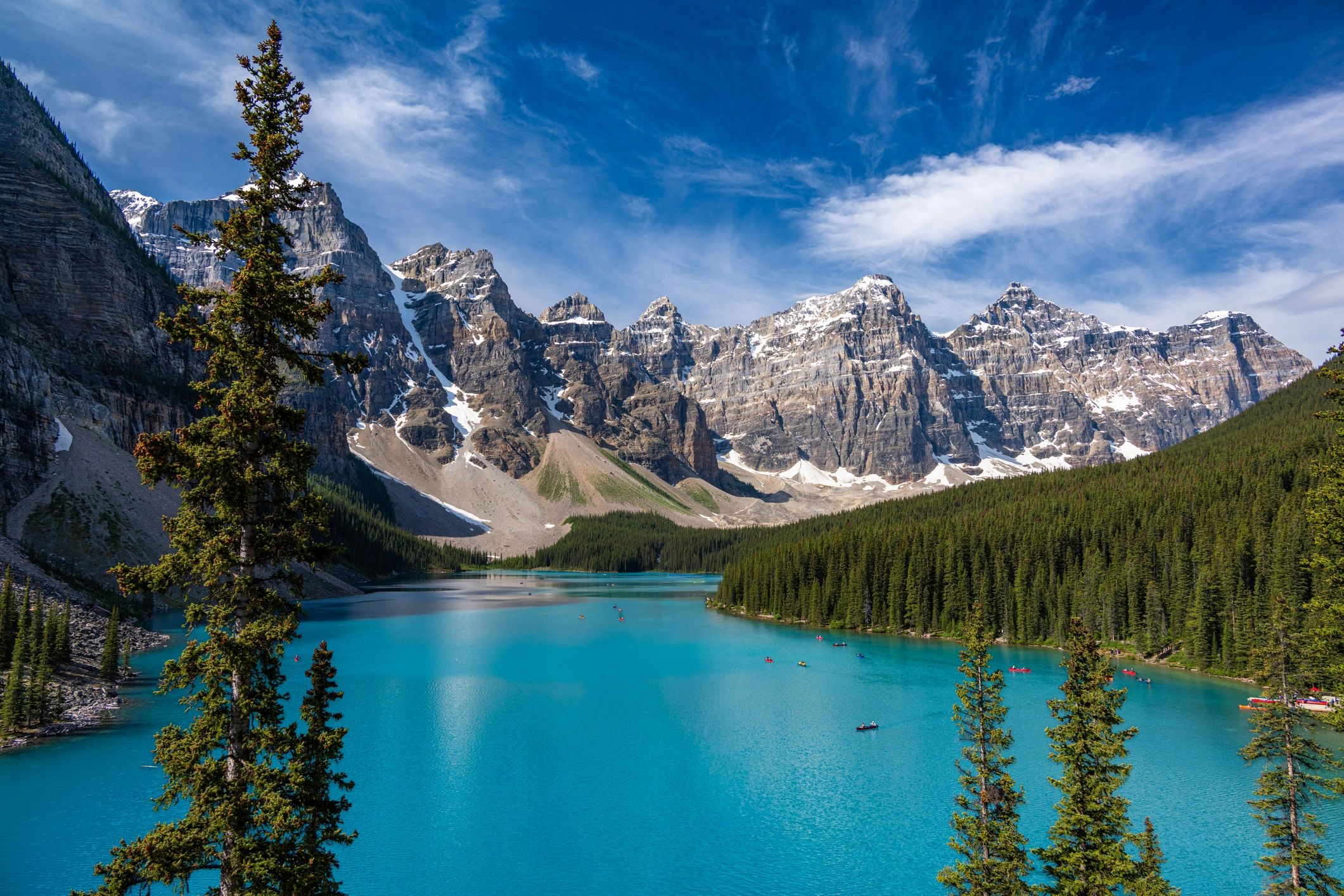 Turquoise lake with snow-capped mountains and pine trees under a blue sky.