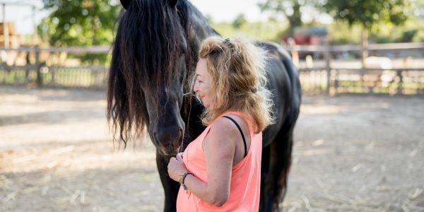 Woman smiling at a black horse in a sunny outdoor setting.