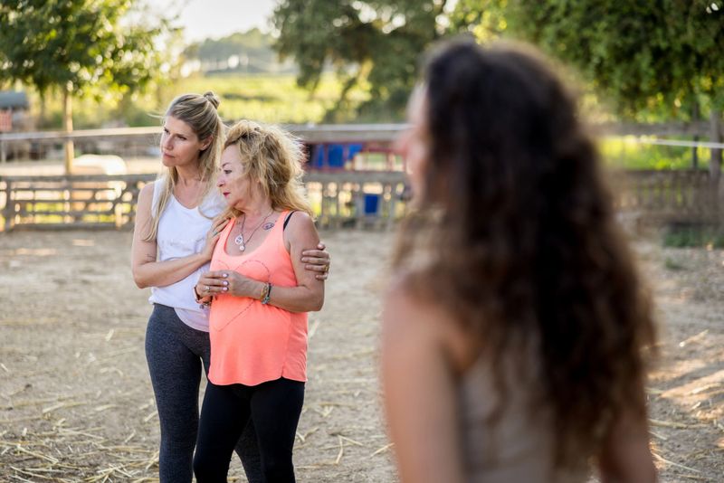 Women supporting each other during equine-assisted therapy, sharing emotional healing moments near horses in tranquil outdoor environment, connecting through therapeutic interaction
