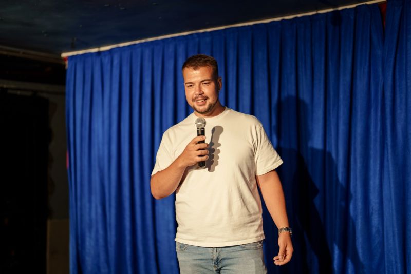 Young man standing in front of a blue curtain with a microphone, smiling and addressing the audience. Concept of stand-up comedy, public speaking, or live performance.