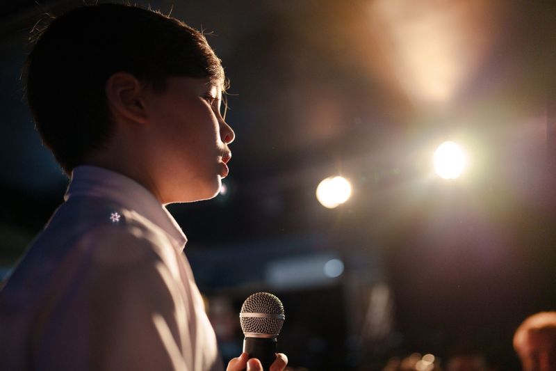Young boy standing on stage holding a microphone, speaking confidently to an audience. Blue curtain background, people watching in foreground. Concept of school performance, public speaking, or talent show.