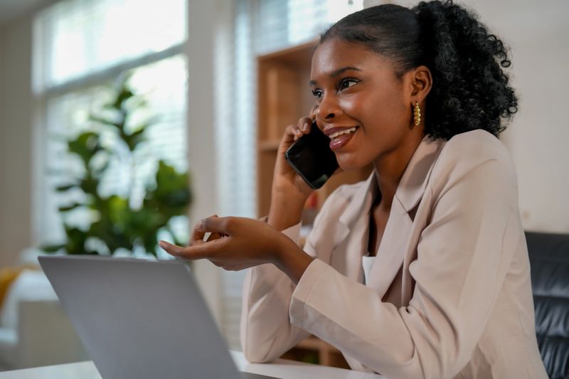 Woman engages in lively conversation on her smartphone while seated at desk with laptop. This scene highlights connection between technology and creative collaboration in modern workspace