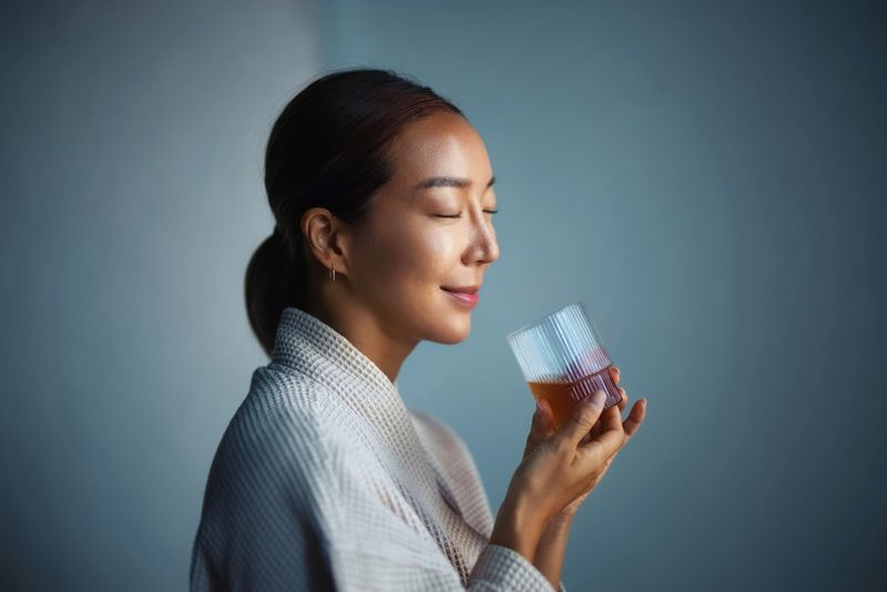 A serene woman with closed eyes holding a beverage glass, portraying relaxation and tranquility in a minimalistic setting, emphasizing mindfulness and enjoyment of peaceful moments.