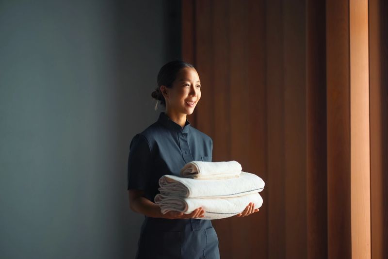 A smiling professional holding neatly folded white towels in a modern wood-paneled environment, symbolizing hospitality and cleanliness.