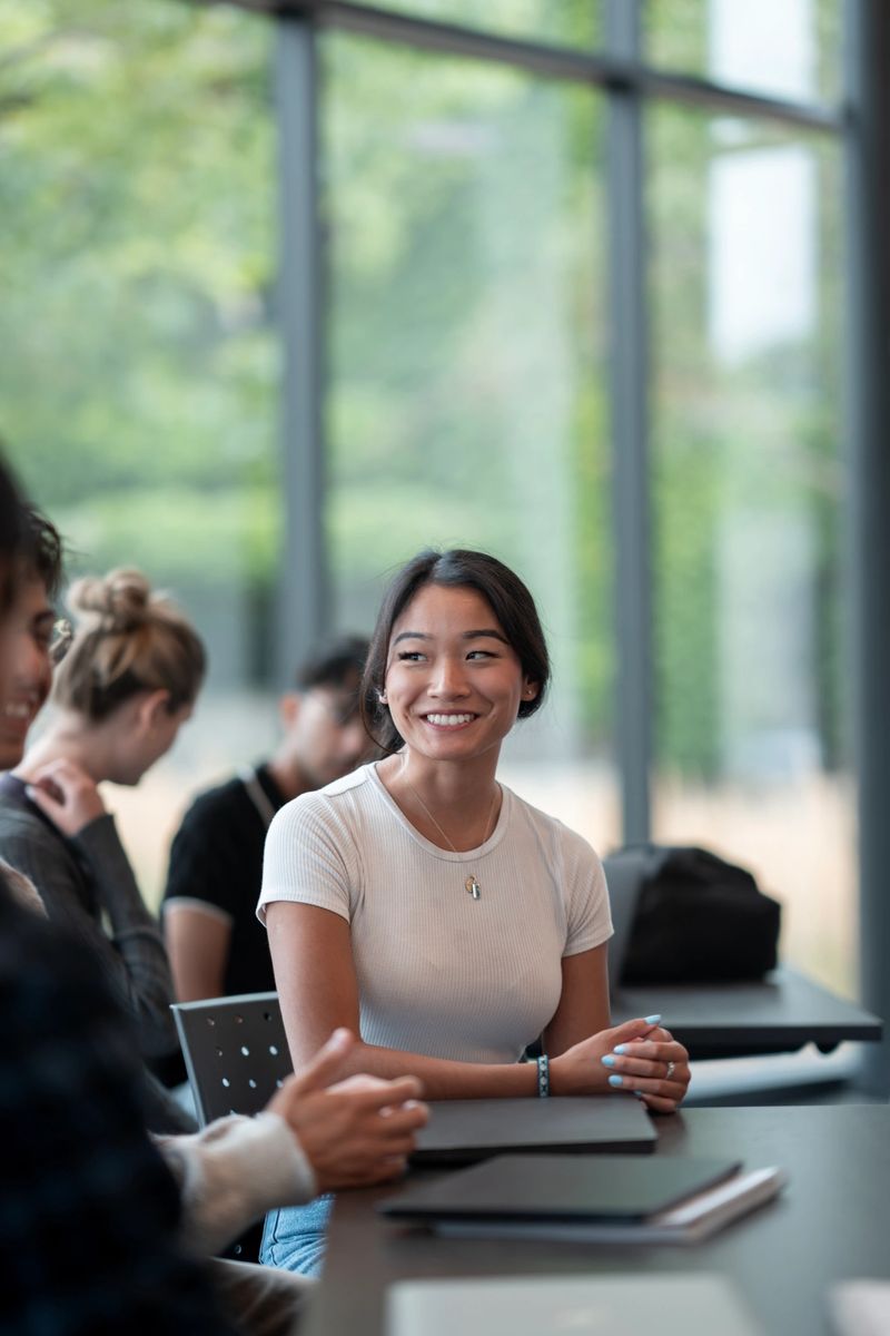 A cheerful young woman wearing casual clothes engages in a modern study group session inside a well-lit library, surrounded by fellow students and lush green scenery visible through large windows.