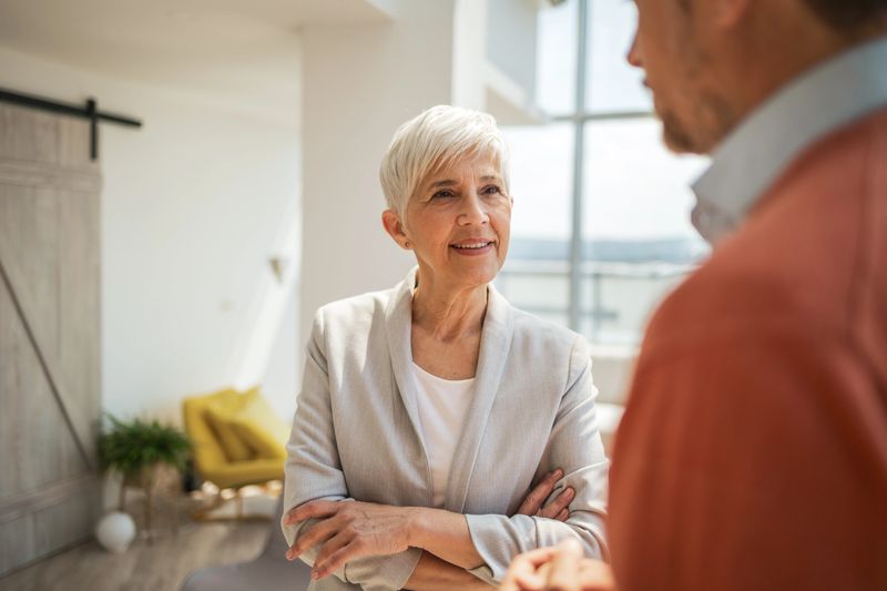 Elderly professional woman standing thoughtfully, speaking to a colleague in a spacious, brightly lit office. The space and interaction evoke themes of communication, experience, and professional collaboration.
