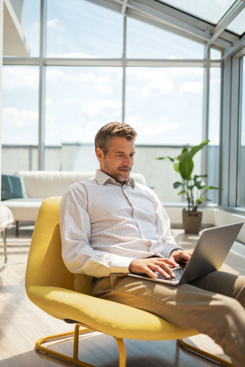 A man is seated on a yellow chair in a contemporary setting with large windows, working on his laptop and appearing focused. The atmosphere is bright, serene, and conducive to productivity.