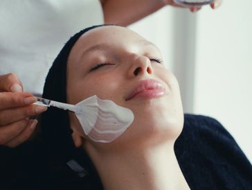 A woman enjoys a facial treatment with a creamy mask applied to her cheek.