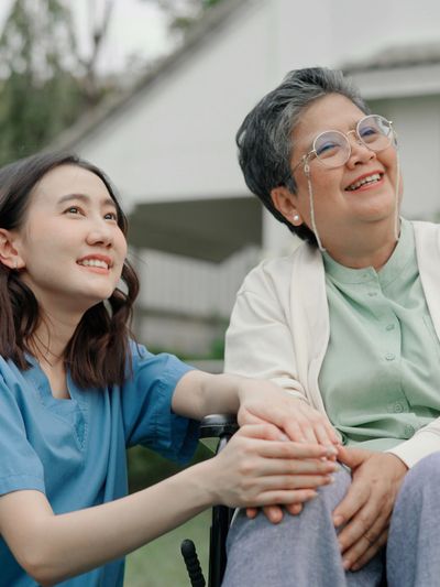 A young caregiver holding hands with a smiling elderly woman outdoors.