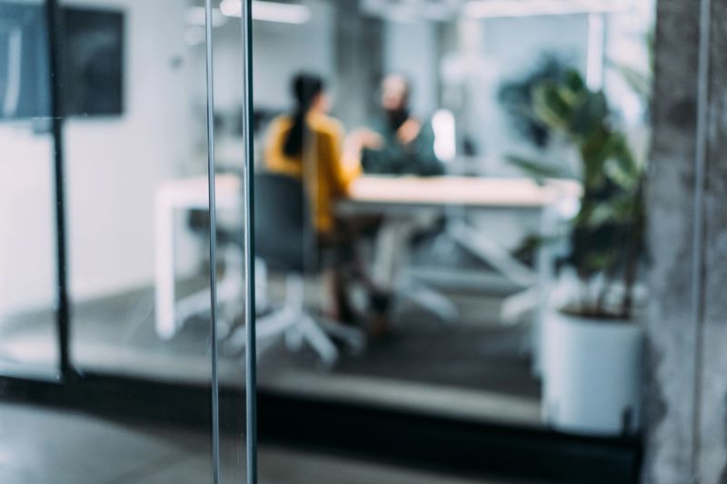 Defocused shot of two colleagues having a discussion in modern office. Confident business people working together in the office. Creative business persons discussing new project and sharing ideas in the workplace. The view is through glass.