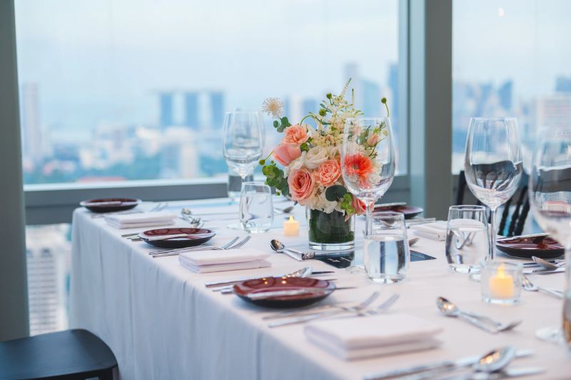 A beautifully set dining table with elegant glassware, silverware, and a floral centerpiece overlooking an urban cityscape. The ambiance is enhanced by soft candlelight, presenting a sophisticated setting for a formal gathering or special event.