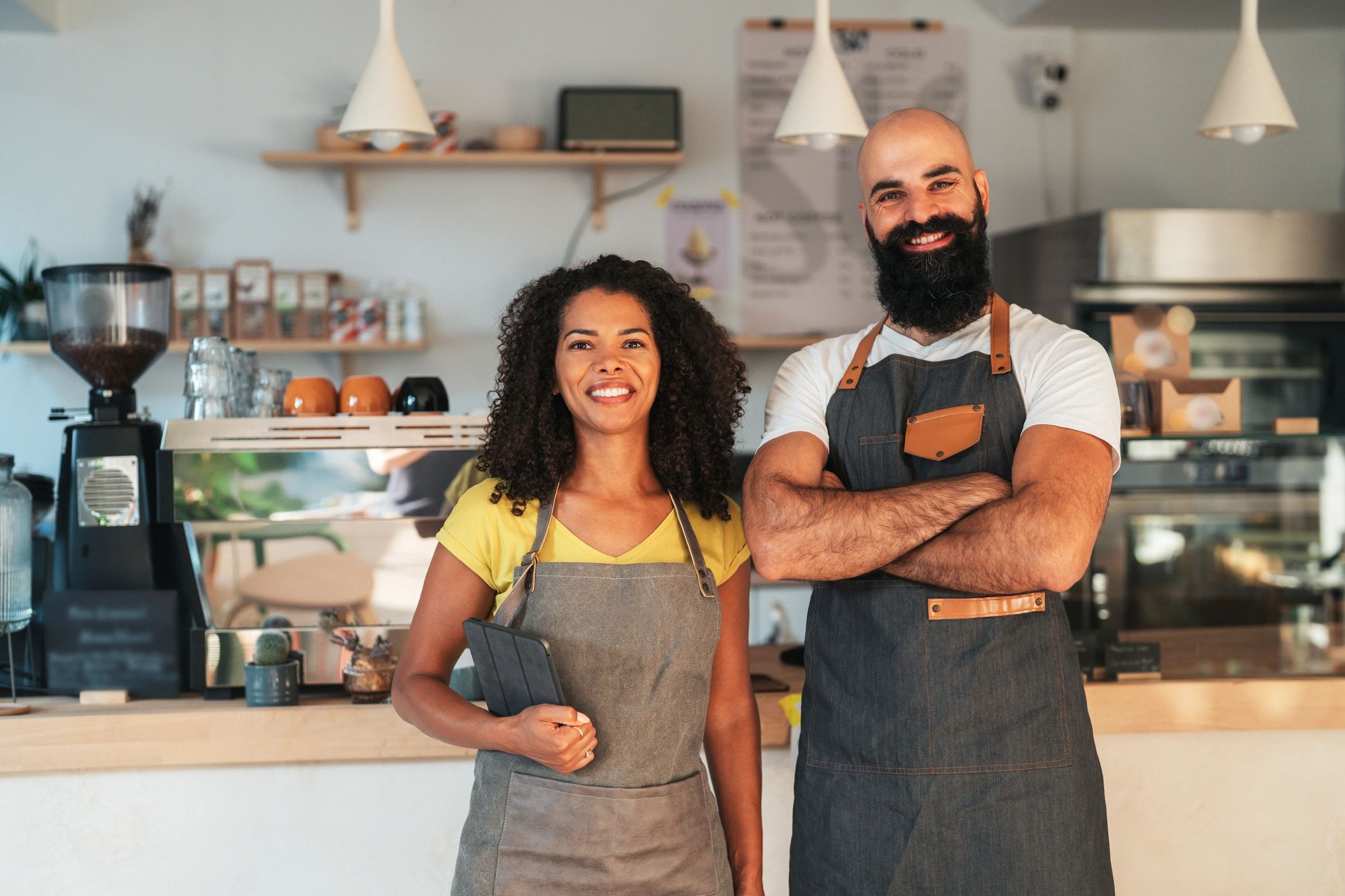 Two smiling baristas in aprons standing confidently in a coffee shop.