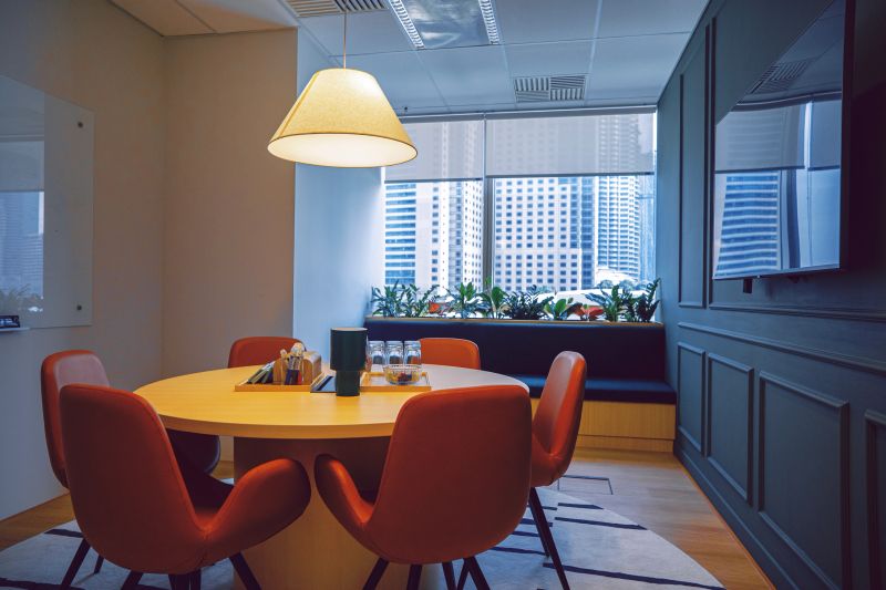 Contemporary office meeting room with round wooden table, orange chairs, and city skyscraper view through large windows. Ideal for concepts of business collaboration and professional workspaces. Kuala Lumpur.