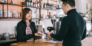 A barista smiling as she takes a payment from a customer at a coffee shop.