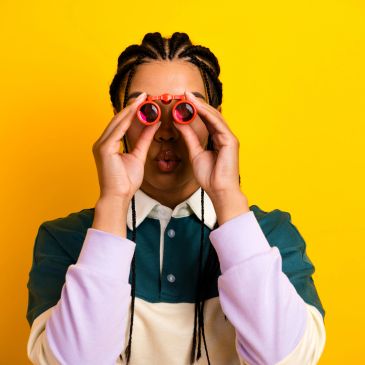 Person with braided hair looking through red binoculars against a yellow background.