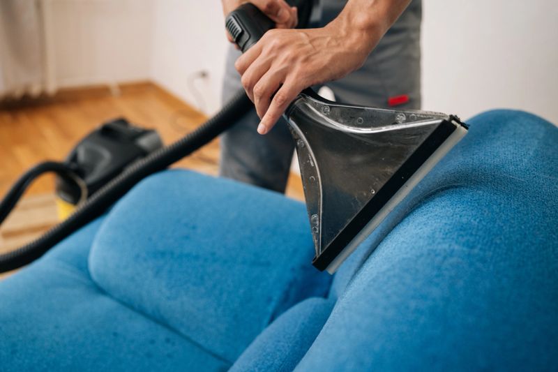 Close-up cropped shot of unrecognizable professional cleaning service worker removing dirt and stains from blue upholstered sofa using extraction washing vacuum cleaner in living room.