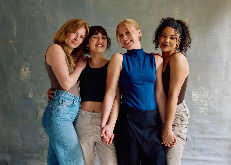 Group of diverse stylish women in their 20s standing close, smiling at camera, representing togetherness, youth culture, and positive emotion in a studio portrait.