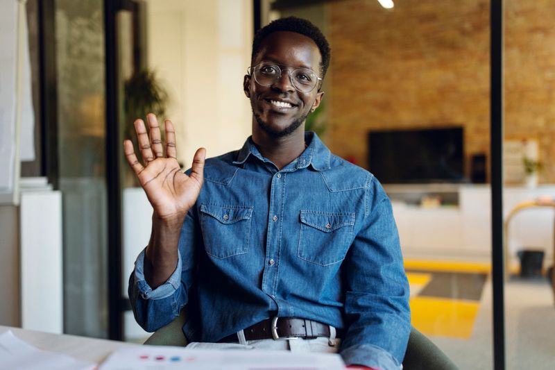 Smiling employee sitting indoors in a casual setting, waving and expressing a friendly demeanor. Bright and welcoming atmosphere with professional yet relaxed ambiance, ideal for subjects related to collaboration and positive interaction in modern workspaces.
