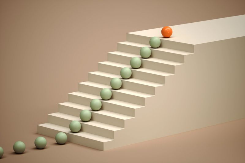 An orange sphere sits at the top of a staircase, distinct from the green spheres below. A 3D illustration representing leadership, uniqueness, and achieving a goal.