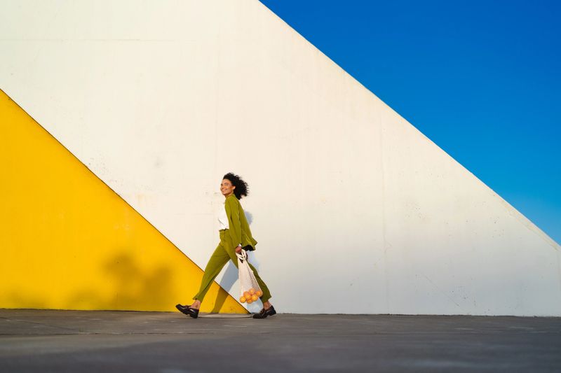 Young woman walking confidently along a vibrant street, carrying a reusable shopping bag, set against a bright, colorful wall